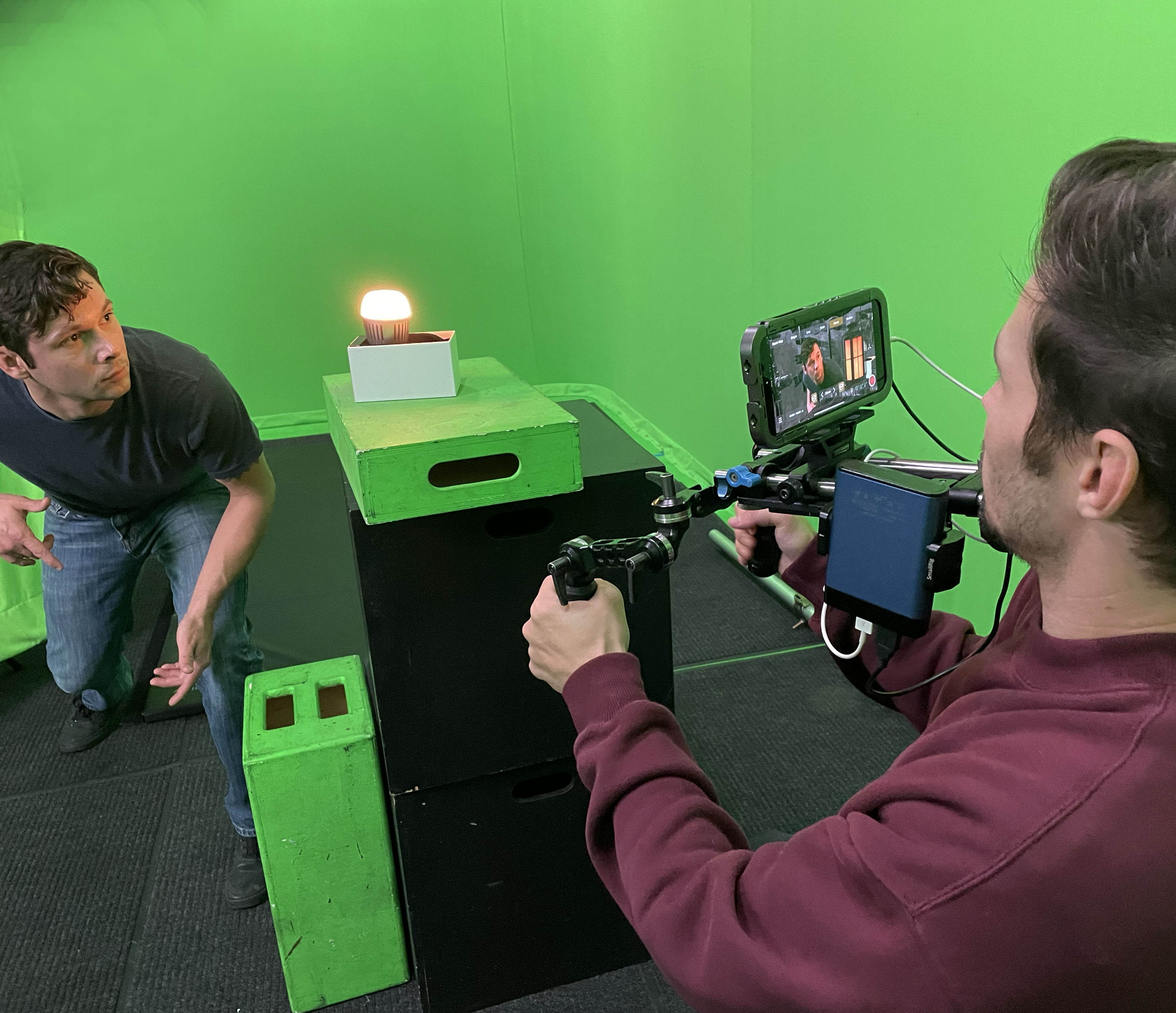 Handheld camera on green screen stage; monitor shows live simulcam composite of performer in a virtual interior